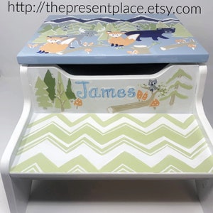 May include: A white wooden step stool with a hand-painted forest scene on top. The top features foxes, a bear, and mountains. The front has the name "James" and a green and white chevron pattern on the step.