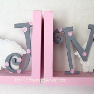 May include: Gray wooden bookends with the letters "JM" in gray with pink polka dots. Each letter has a white lamb with pink accents standing next to it. The bookends are on a pink shelf with the website "http://thepresentplace.etsy.com" printed on it.