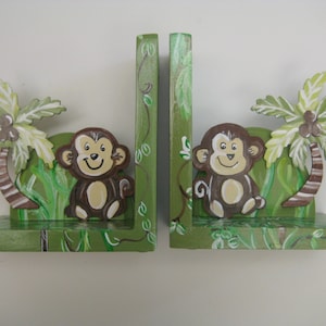 May include: A pair of green wooden bookends with a jungle theme. Each bookend features a smiling monkey sitting in front of a palm tree with brown and white stripes. The bookends are painted with green leaves and vines.