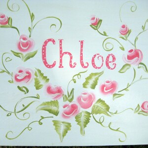 Puede incluir: Un lienzo blanco con el nombre "Chloe" pintado en rosa con lunares blancos. El nombre está rodeado de rosas rosas y hojas verdes.