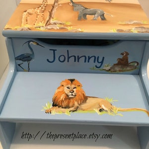 May include: A light blue step stool with hand-painted safari animals. The top features a giraffe, elephant, and monkey. The front has the name "Johnny" and a lion. The website address is at the bottom.