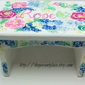 May include: White wooden step stool hand-painted with pink roses, blue hydrangeas, and green leaves. The name "Rose" is written in pink script. The legs and edges are also decorated with floral designs. The Etsy shop URL is visible.