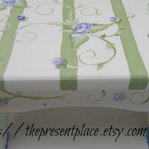 May include: A white painted wooden stool with green stripes and a hand-painted floral design featuring purple flowers and green leaves. The stool has a website address painted on the bottom: http://thepresentplace.etsy.com.