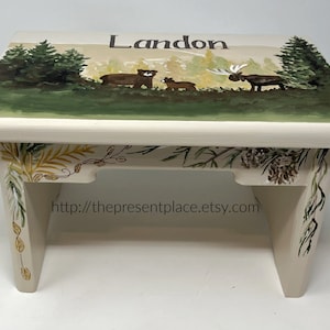 May include: A white wooden step stool with a hand-painted woodland scene featuring a moose, two bears, and pine trees. The stool is decorated with a leafy border and the name "Landon" is painted on the top. The website address "http://thepresentplace.etsy.com" is painted on the front of the stool.