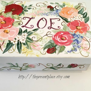 Puede incluir: Una caja de madera blanca con un diseño floral. La parte superior está adornada con flores rojas, rosas y naranjas, hojas verdes y el nombre "ZOE" en escritura púrpura. La base de la caja presenta un patrón floral a juego.