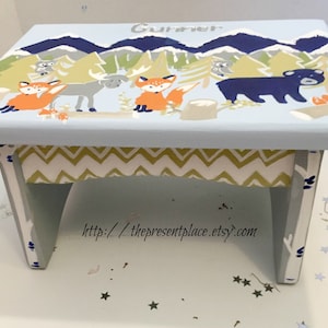 May include: A light blue wooden step stool with a whimsical woodland scene painted on the top. The scene includes a fox, a moose, an owl, and a bear. The stool has a chevron patterned fabric lining the inside. The text "Gummer" is painted on the top of the stool.