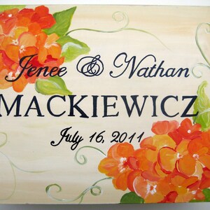 Op de afbeelding: Handgeschilderd houten bord met de namen "Jenee & Nathan MACKIEWICZ" en de datum "16 juli 2011". Het bord is versierd met oranje en gele bloemen en groene bladeren.