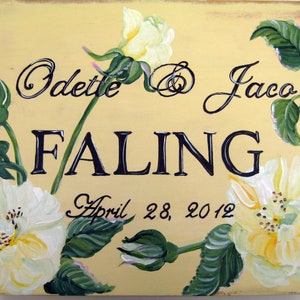 Op de afbeelding: Handgeschilderd houten bord met een lichtgele achtergrond. Het bord toont witte rozen en groene bladeren. De tekst bevat de namen "Odette & Jaco" en "Faling", samen met de datum "28 april 2012".