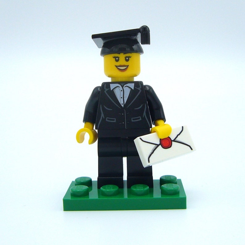 May include: A black graduation cap and gown Lego minifigure holding a white envelope with a red seal. The minifigure is standing on a green base.