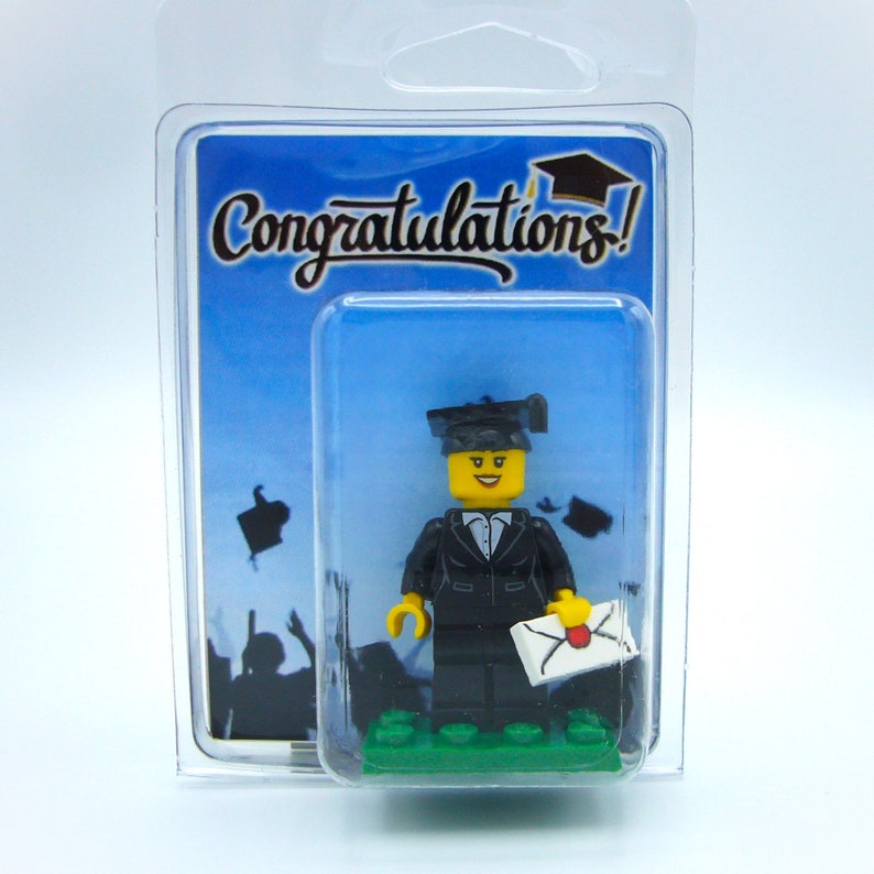 May include: A Lego minifigure wearing a black graduation gown and cap, holding a white envelope with a red seal. The minifigure is standing on a green base. The background is a blue sky with black silhouettes of people celebrating.