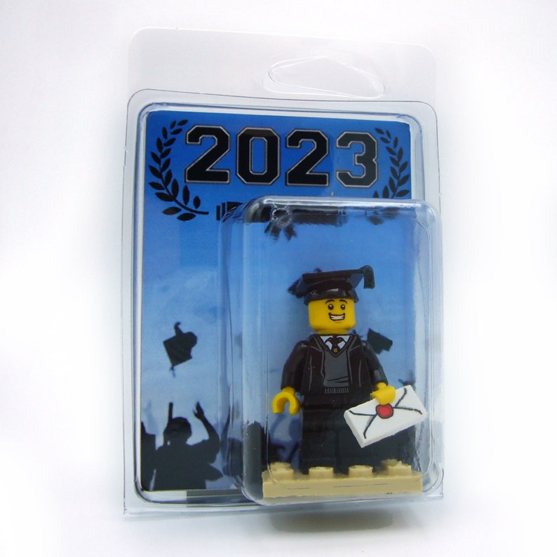 May include: A Lego minifigure dressed in a black graduation gown and cap, holding a diploma. The minifigure is standing on a small platform with the year 2023 printed on the background.