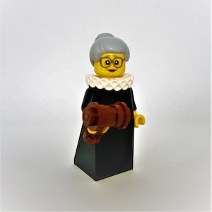 May include: A Lego minifigure of a woman wearing a black robe, a white collar, and glasses. She has gray hair and is holding a brown gavel.