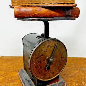 May include: An antique black metal scale with a brass face and a dial that reads up to 23 pounds. Two leather-bound books are on the scale platform.