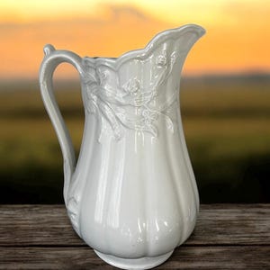 May include: White ceramic pitcher with a fluted design and floral embossed details. The pitcher has a wide mouth and a curved handle.