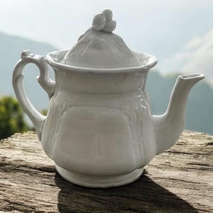 May include: A white ceramic teapot with a lid and a curved handle. The lid is topped with three small, round decorative elements. The teapot is sitting on a wooden surface, with a mountain range in the background.