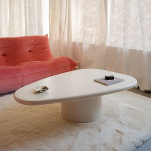 Könnte beinhalten: Ein weißer, ovaler Couchtisch mit zylindrischem Fuß steht auf einem cremefarbenen Teppich. Im Hintergrund ist ein rosa Sofa zu sehen. Auf dem Tisch liegen eine kleine Muschel und ein quadratischer Untersetzer mit Räucherstäbchen.