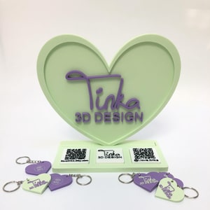 Könnte beinhalten: Ein hellgrüner herzförmiger Präsentationsständer mit dem Text "Tinka 3D DESIGN" in Lila. Mehrere herzförmige Schlüsselanhänger in Hellgrün und Lila werden vor dem Ständer ausgestellt. Die Schlüsselanhänger tragen den Text "Tinka 3D DESIGN".