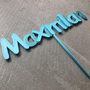 Könnte beinhalten: Ein hellblauer Tortenaufsatz mit dem Namen "Maximilian" in Schreibschrift. Der Aufsatz besteht aus einem 3D-gedruckten Material und hat einen Stab zum Einstecken in einen Kuchen. Der Hintergrund ist eine graue Oberfläche.