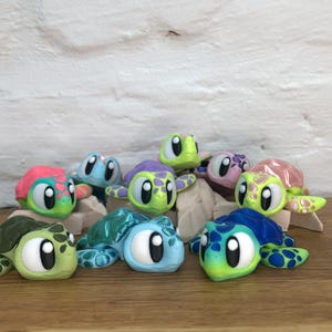 Könnte beinhalten: Eine Sammlung farbenfroher Schildkrötenfiguren. Jede Schildkröte hat ein einzigartiges Schalenmuster und eine einzigartige Farbe, darunter Grün, Blau, Rosa und Lila. Die Schildkröten haben große, ausdrucksstarke Augen und sind auf einer Holzoberfläche angeordnet.