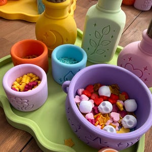 Könnte beinhalten: Eine Sammlung bunter Silikon-Zaubertrankflaschen, kleiner Becher und einer großen Schale auf einem hellgrünen Tablett. Die Gegenstände haben geprägte Motive wie Monde, Blätter, Herzen und Blumen. Die Schale enthält kleine Totenkopf-, Herz- und Sternformen.