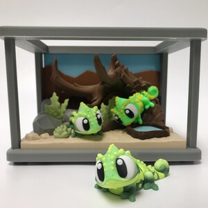 Könnte beinhalten: Eine Miniatur-Terrarienszene mit drei Cartoon-Chamäleons in Grün- und Gelbtönen. Die Szene umfasst einen Hintergrund mit blauem Himmel, braunen Ästen und einem kleinen Wasserbecken. Das Terrarium ist in einem grauen Rahmen eingeschlossen.