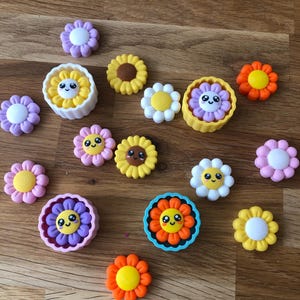 Könnte beinhalten: Eine Sammlung kleiner blumenförmiger Silikonformen und passender blumenförmiger Einsätze. Die Teile zeigen lächelnde Gesichter mit großen Augen und sind in den Farben Gelb, Orange, Lila, Rosa, Weiß und Hellblau gehalten.