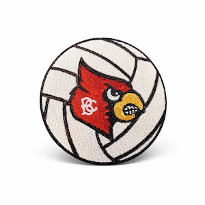 K&ouml;nnte beinhalten: Ein runder Aufn&auml;her mit einem Volleyball-Design und einem roten Kardinal-Maskottchen. Der Kardinal hat einen gelben Schnabel und ist vor einem wei&szlig;-schwarzen Volleyballmuster platziert. Der Aufn&auml;her ist wahrscheinlich f&uuml;r Sportfans.