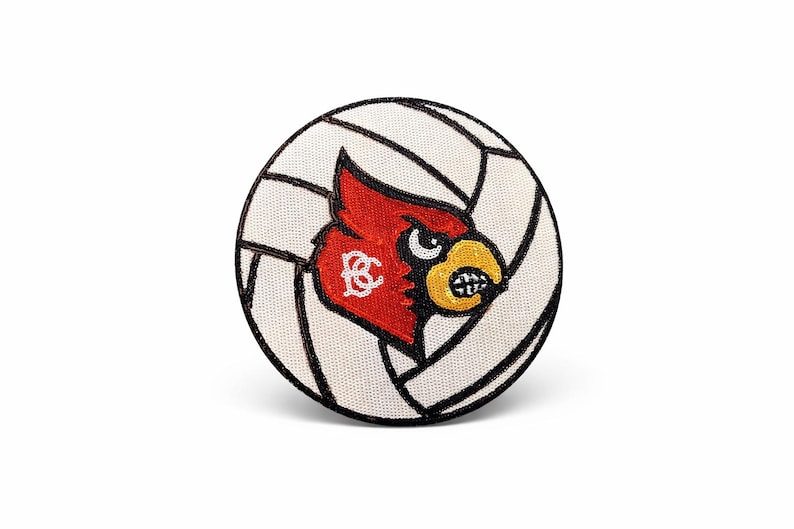 K&ouml;nnte beinhalten: Ein runder Aufn&auml;her mit einem Volleyball-Design und einem roten Kardinal-Maskottchen. Der Kardinal hat einen gelben Schnabel und ist vor einem wei&szlig;-schwarzen Volleyballmuster platziert. Der Aufn&auml;her ist wahrscheinlich f&uuml;r Sportfans.