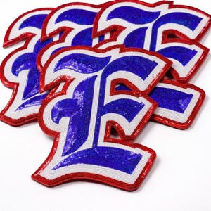 K&ouml;nnte beinhalten: Eine Ansammlung von Buchstaben-Patches in Form des Buchstabens "F". Die Patches haben eine blaue Paillettenmitte, einen wei&szlig;en Rand und eine rote Umrandung. Die Patches sind leicht &uuml;berlappend auf wei&szlig;em Hintergrund angeordnet. H&ouml;he ca. 10 cm.