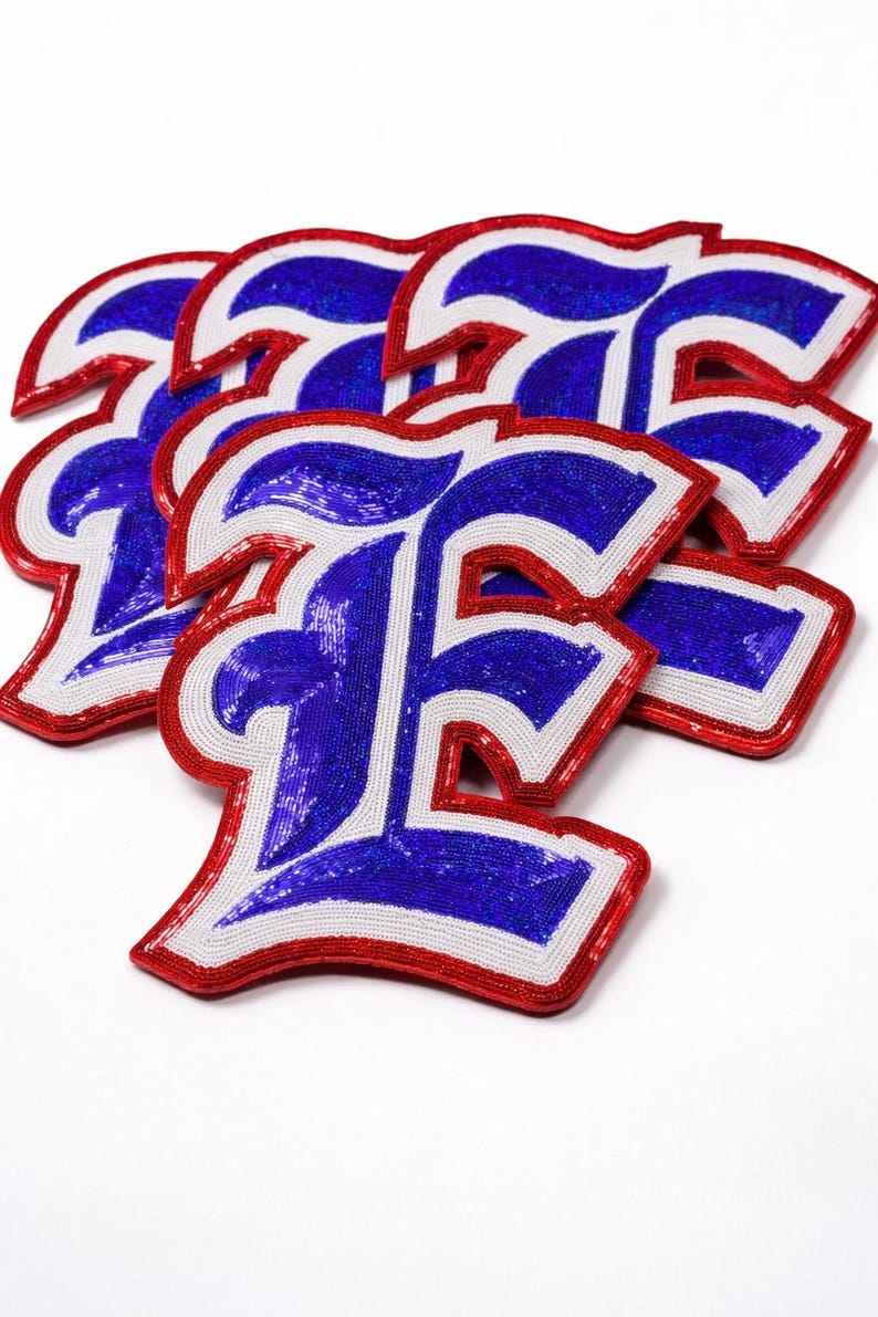 K&ouml;nnte beinhalten: Eine Ansammlung von Buchstaben-Patches in Form des Buchstabens "F". Die Patches haben eine blaue Paillettenmitte, einen wei&szlig;en Rand und eine rote Umrandung. Die Patches sind leicht &uuml;berlappend auf wei&szlig;em Hintergrund angeordnet. H&ouml;he ca. 10 cm.
