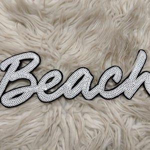 K&ouml;nnte beinhalten: Ein wei&szlig;er Aufn&auml;her mit dem Wort "Beach" in Schreibschrift, mit schwarzem Rand und verziert mit kleinen, gl&auml;nzenden, silberfarbenen Perlen. Der Aufn&auml;her befindet sich auf einem flauschigen, hellfarbigen Hintergrund.