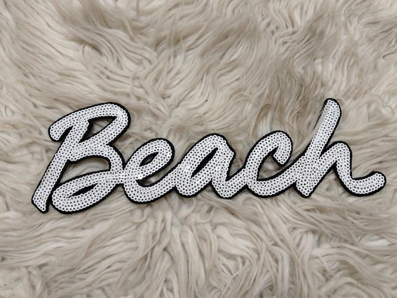 K&ouml;nnte beinhalten: Ein wei&szlig;er Aufn&auml;her mit dem Wort "Beach" in Schreibschrift, mit schwarzem Rand und verziert mit kleinen, gl&auml;nzenden, silberfarbenen Perlen. Der Aufn&auml;her befindet sich auf einem flauschigen, hellfarbigen Hintergrund.