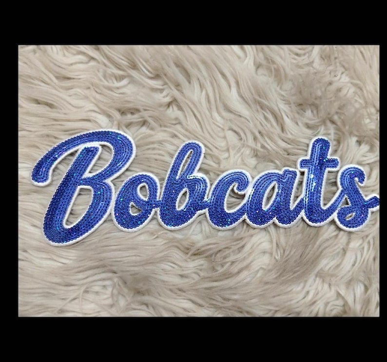 K&ouml;nnte beinhalten: Blauer Pailletten-Schriftzug "Bobcats" mit wei&szlig;em Rand. Das Wort ist in einer Schreibschrift dargestellt und befindet sich auf einer flauschigen, hellfarbigen Oberfl&auml;che. Die Pailletten lassen die Buchstaben glitzern.