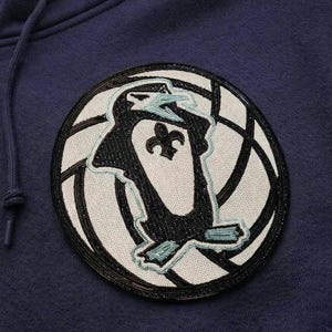 K&ouml;nnte beinhalten: Marineblauer Kapuzenpullover mit einem kreisf&ouml;rmigen Aufn&auml;her mit einem Pinguin-Design. Der Aufn&auml;her hat eine schwarze Perlenkontur und einen wei&szlig;en Hintergrund mit einer schwarz-wei&szlig;en Grafik. Der Pinguin hat ein Fleur-de-Lis-Symbol.