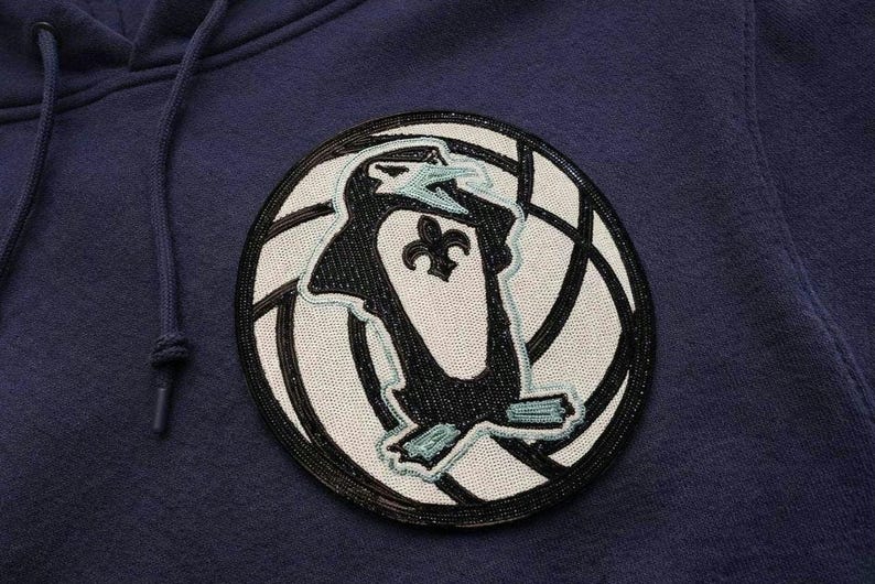 K&ouml;nnte beinhalten: Marineblauer Kapuzenpullover mit einem kreisf&ouml;rmigen Aufn&auml;her mit einem Pinguin-Design. Der Aufn&auml;her hat eine schwarze Perlenkontur und einen wei&szlig;en Hintergrund mit einer schwarz-wei&szlig;en Grafik. Der Pinguin hat ein Fleur-de-Lis-Symbol.