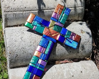 May include: A colorful stained glass cross with a mosaic design. The cross is made of small pieces of glass in shades of blue, green, orange, and pink. The glass is iridescent and has a glittery finish.