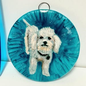 May include: A white dog with a black collar is painted on a round, blue glass ornament. The ornament has a silver ring at the top for hanging.