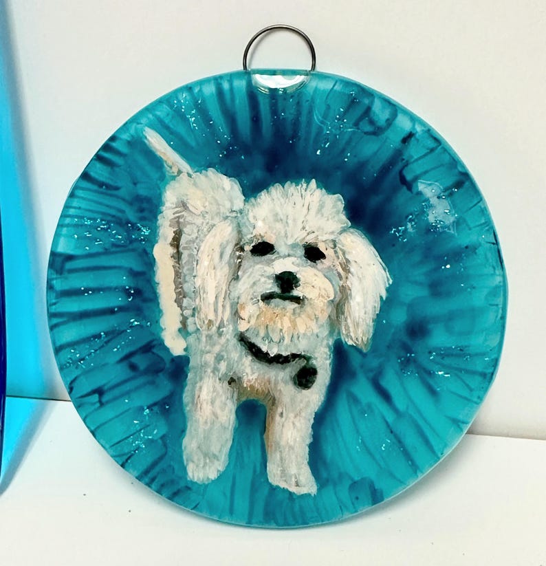 May include: A white dog with a black collar is painted on a round, blue glass ornament. The ornament has a silver ring at the top for hanging.