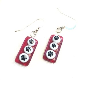 May include: A pair of red glass earrings with three white paw prints on each earring. The earrings have silver hooks.