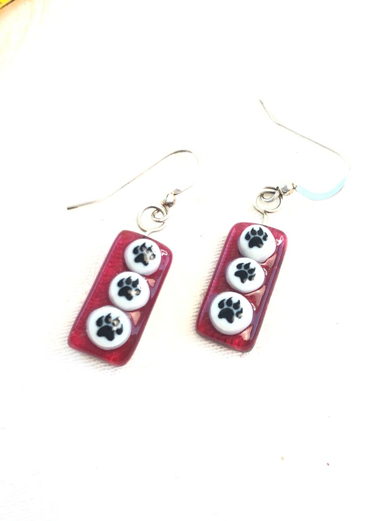 May include: A pair of red glass earrings with three white paw prints on each earring. The earrings have silver hooks.