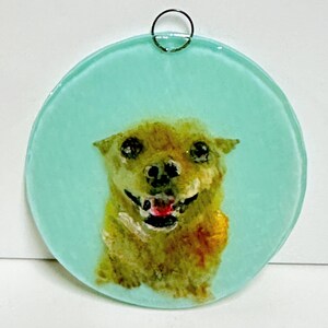 May include: A round glass ornament with a light blue background and a painted image of a brown dog with a red tongue sticking out. The dog is looking directly at the viewer.