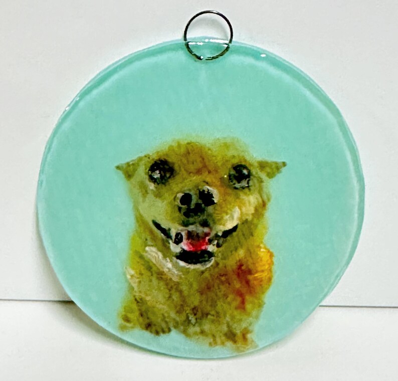 May include: A round glass ornament with a light blue background and a painted image of a brown dog with a red tongue sticking out. The dog is looking directly at the viewer.