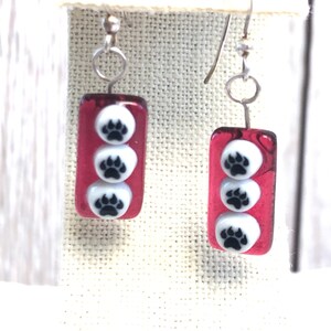 May include: A pair of red glass earrings with three white paw prints on each earring. The earrings have silver hooks.