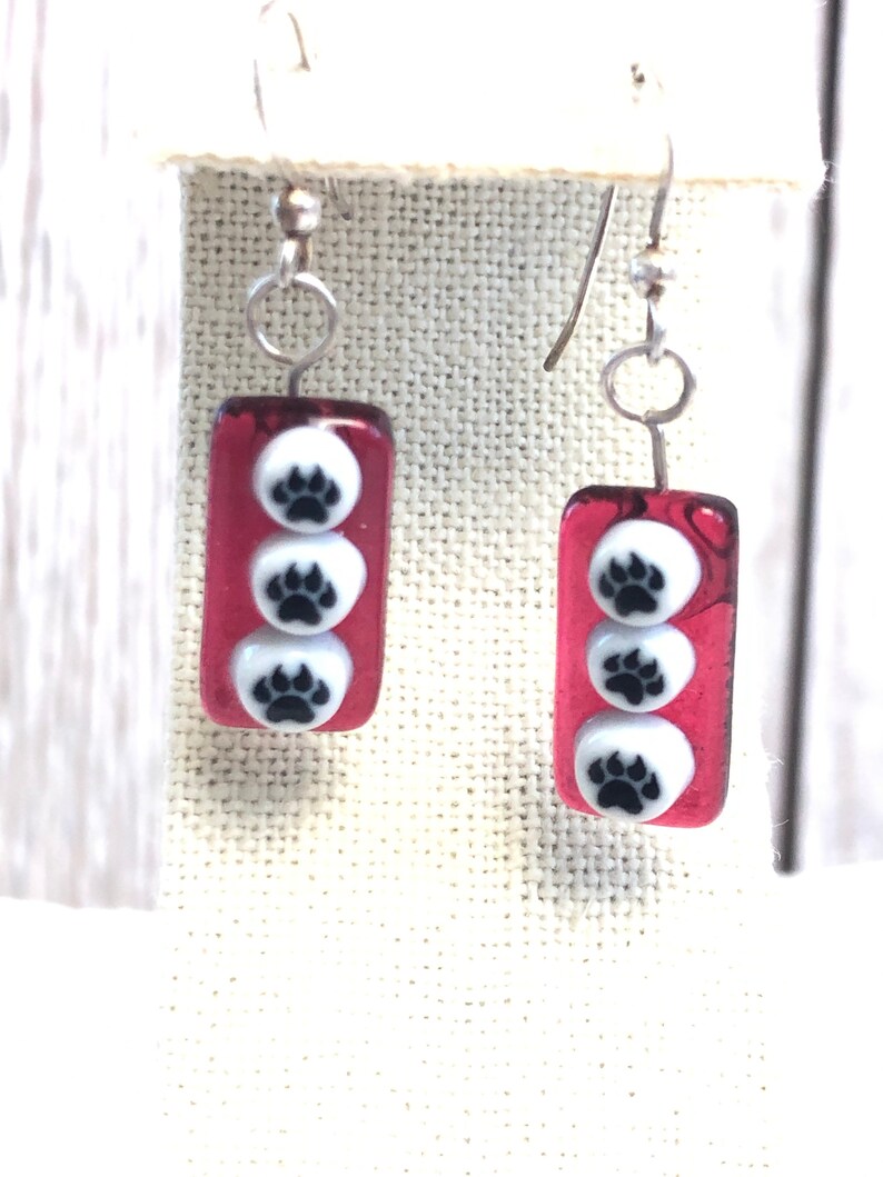 May include: A pair of red glass earrings with three white paw prints on each earring. The earrings have silver hooks.