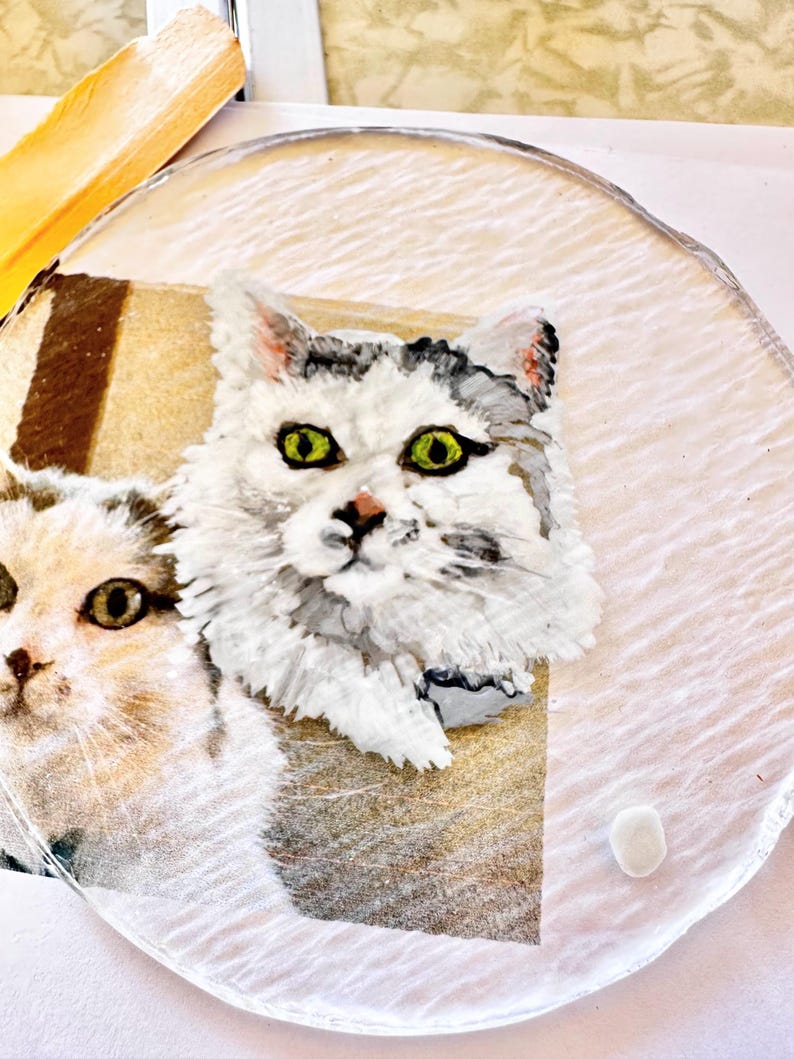 May include: A close-up of a white, gray, and black cat with green eyes. The cat is painted on a round, clear glass surface.