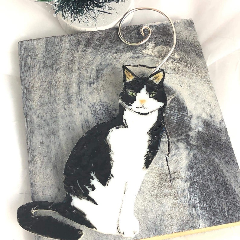 May include: Black and white cat Christmas ornament with a wire hanger. The cat is painted in a realistic style and has green eyes.