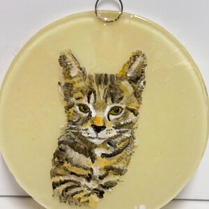 May include: A round glass ornament with a painted image of a tabby cat. The cat is looking directly at the viewer with a light yellow background.