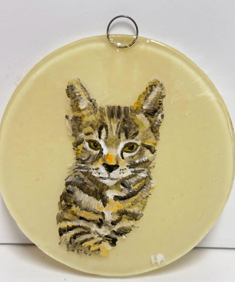 May include: A round glass ornament with a painted image of a tabby cat. The cat is looking directly at the viewer with a light yellow background.