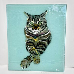 May include: A painting of a tabby cat with green eyes on a light blue background. The cat is sitting with its paws crossed and has a purple collar with a blue jewel.