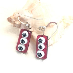 May include: Red glass dangle earrings with silver hooks and three white circles with black paw prints on each earring.